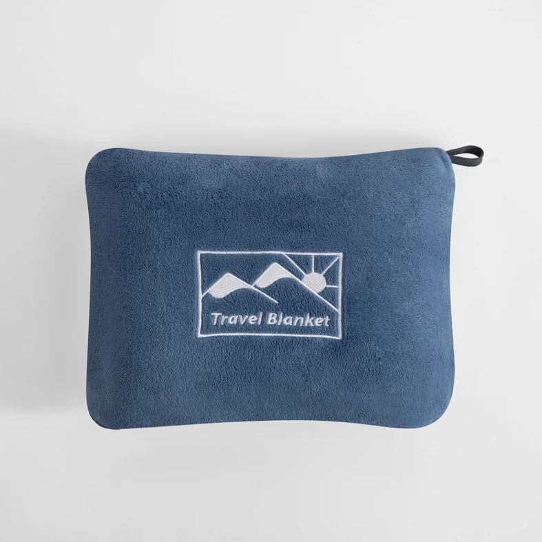 Niebieski koc podróżny złożony do pokrowca o tym samym kolorze. Na pokrowcu logo przedstawiające słońce wychodzące zza gór oraz napis Travel Blanket. U jednego z rogów pokrowca pętelka-uchwyt.
