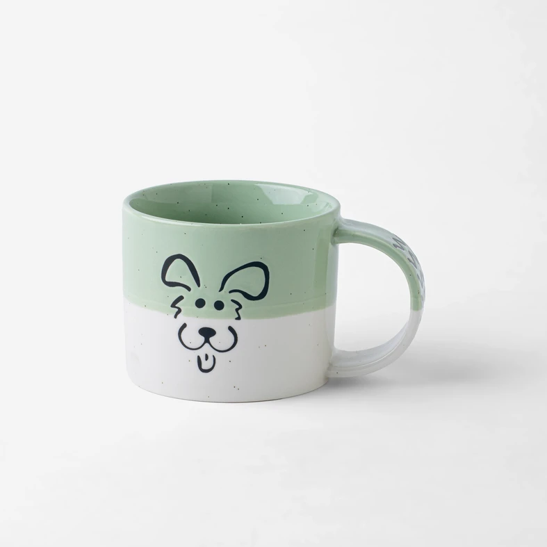 Porcelanowy kubek, w połowie jasnozielony, w połowie biały. Na ściance kontur radosnego psiego pyszczka. Kubek ma szerokie ucho.