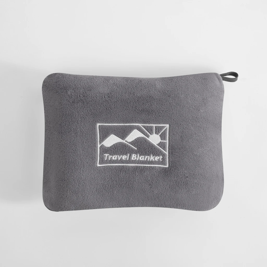 Szary koc podróżny złożony do pokrowca o tym samym kolorze. Na pokrowcu logo przedstawiające słońce wychodzące zza gór oraz napis Travel Blanket. U jednego z rogów pokrowca pętelka-uchwyt.