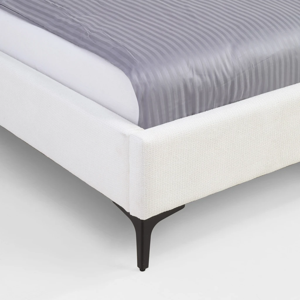 Zbliżenie na kant łóżka. Widoczna struktura białej tkaniny tapicerskiej i czarna nóżka.