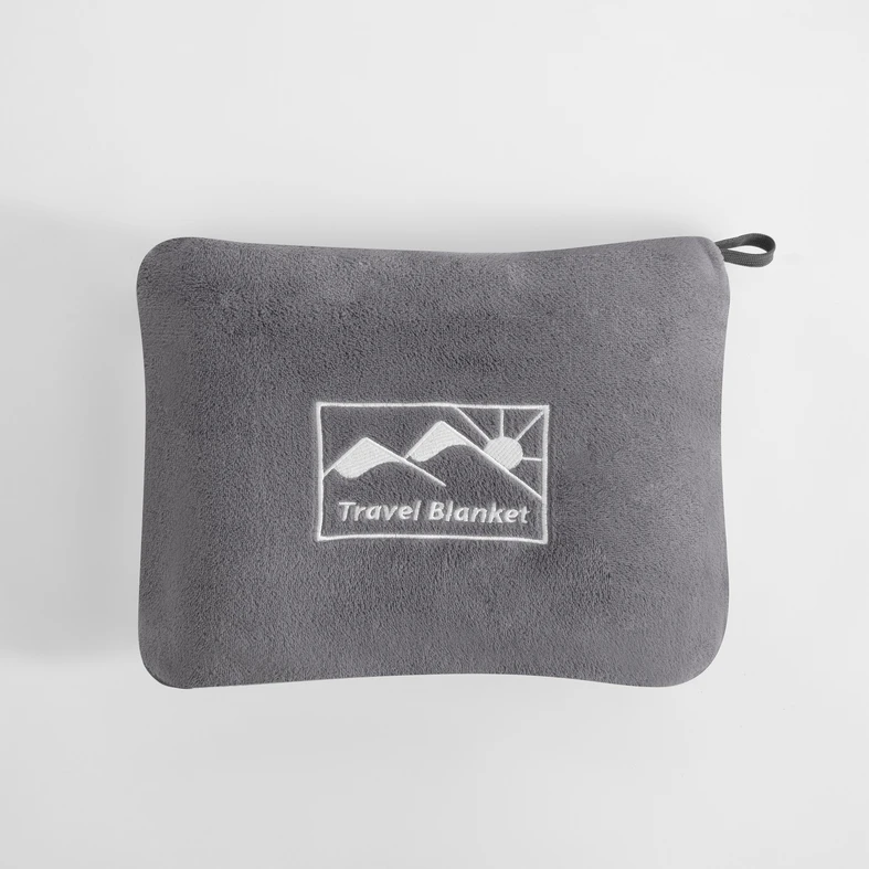 Szary koc podróżny złożony do pokrowca o tym samym kolorze. Na pokrowcu logo przedstawiające słońce wychodzące zza gór oraz napis Travel Blanket. U jednego z rogów pokrowca pętelka-uchwyt.