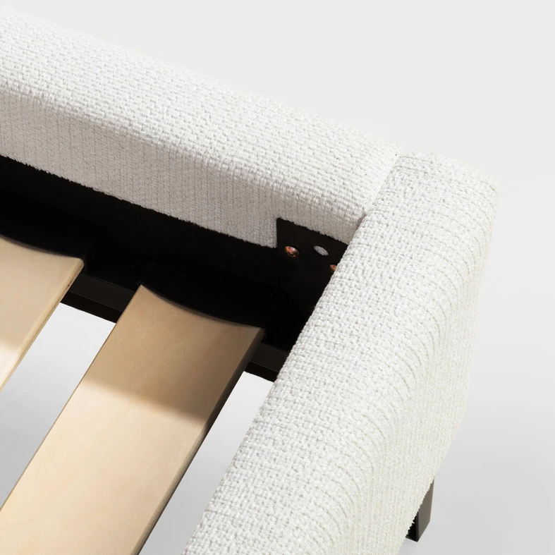 Zbliżenie na róg łóżka, widoczne szczebelki stelaża i fragment czarnej ramy. Boki obite białą tkaniną.