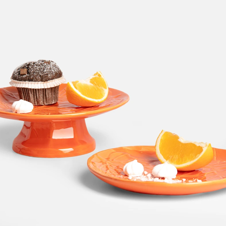 Dwie pomarańczowe patery z wzorem liści, na jednej muffin, na drugiej kawałki pomarańczy i bezy.