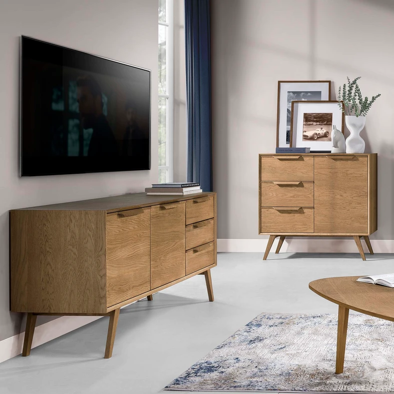 Zdjęcie aranżacyjne, komoda w stylu retro ukazana pod kątem, stoi w salonie utrzymanym w jasnych stonowanych barwach. Na blacie książki. Nad komodą na ścianie zawieszony telewizor.