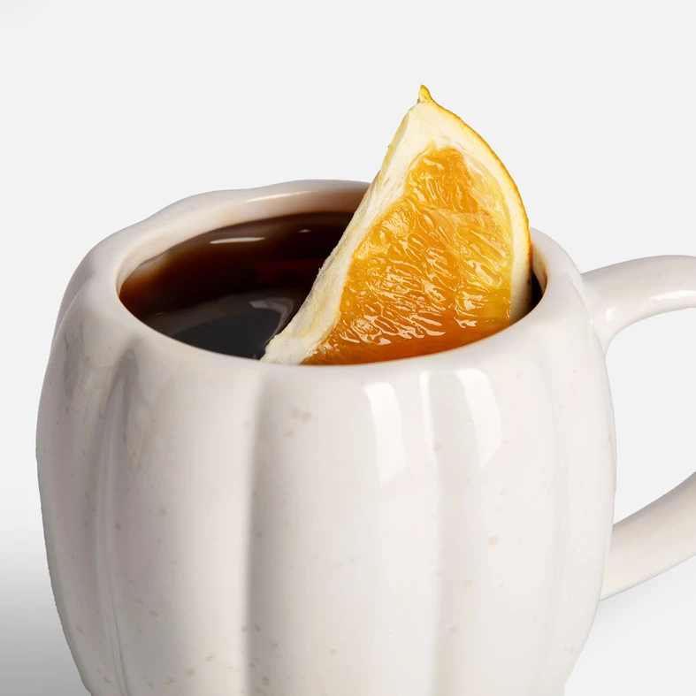 Zbliżenie na biały, ceramiczny kubek w kształcie dyni. W środku napój z pomarańczą