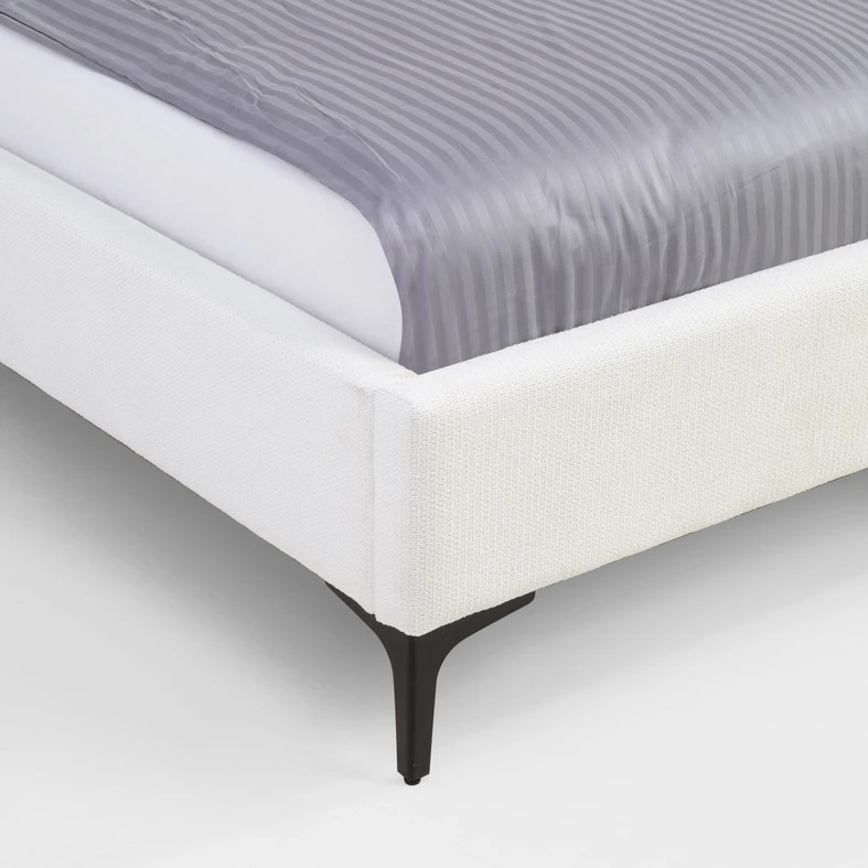 Zbliżenie na kant łóżka. Widoczna struktura białej tkaniny tapicerskiej i czarna nóżka.