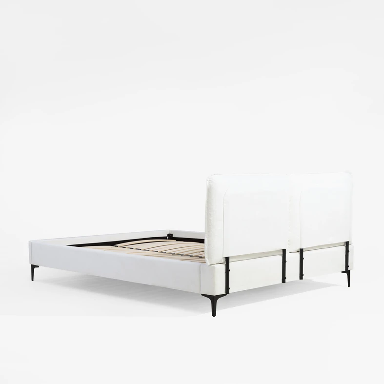 Łózko z białą tkaniną tapicerską. Ma tapicerowany wysoki zagłówek i boki. Widoczne smukłe elementy czarnej ramy.