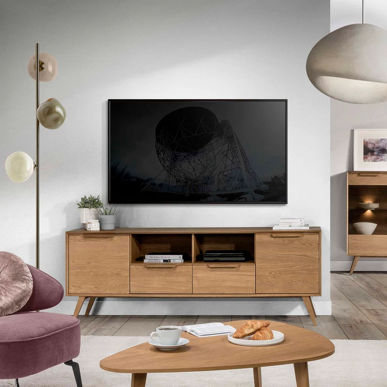 Zdjęcie aranżacyjne, komoda RTV w stylu retro stoi w salonie utrzymanym w jasnych stonowanych barwach. Na blacie rośliny w doniczkach i książki. Ponad komodą zawieszony telewizor.